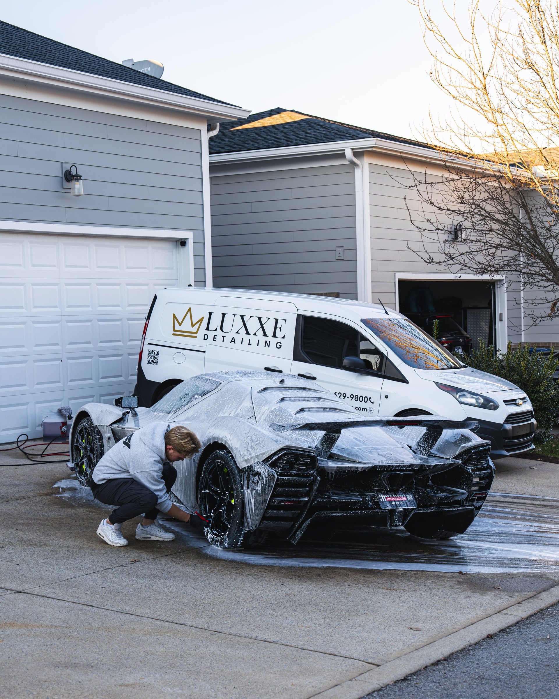 Worker wrapping luxury black sports car with protective film on driveway, white van with branding in background