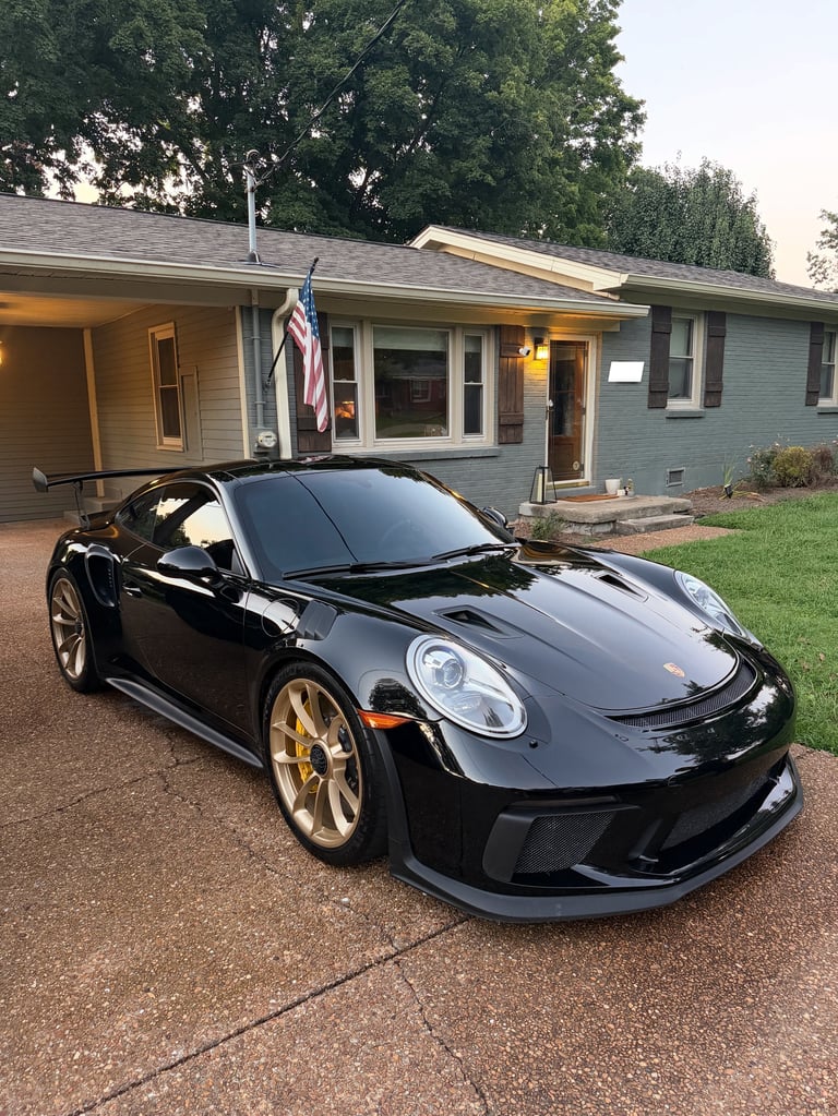 Black Porsche 911 GT3 with gold wheels parked on driveway in front of blue house