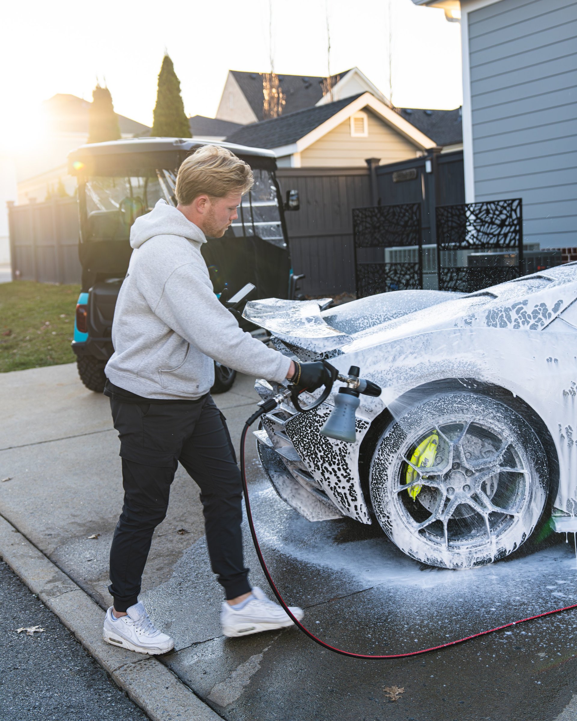 Man in gray hoodie washing white car with foam and hose in residential driveway during sunset